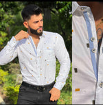 A man wearing a long sleeve printed shirt in white with a geometric pattern, with black trouser standing outdoors.