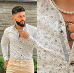 A man wearing a long sleeve printed shirt in white with gray geometric patterns, with beige trouser standing outdoors.